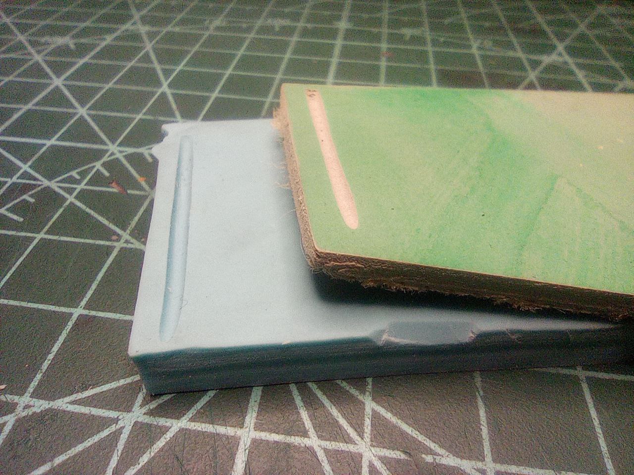 Foto a dos trozos de planchas de linograbado puestas sobre una superficie de trabajo verde. Una es de goma azul, y un surco hecho muestra el mismo color en toda su composición. Encima está un trozo de linóleo que tiene la superficie pintada de verde. El surco hecho en esta pieza hace que se vea la diferencia de textura y color con respecto a la superficie.