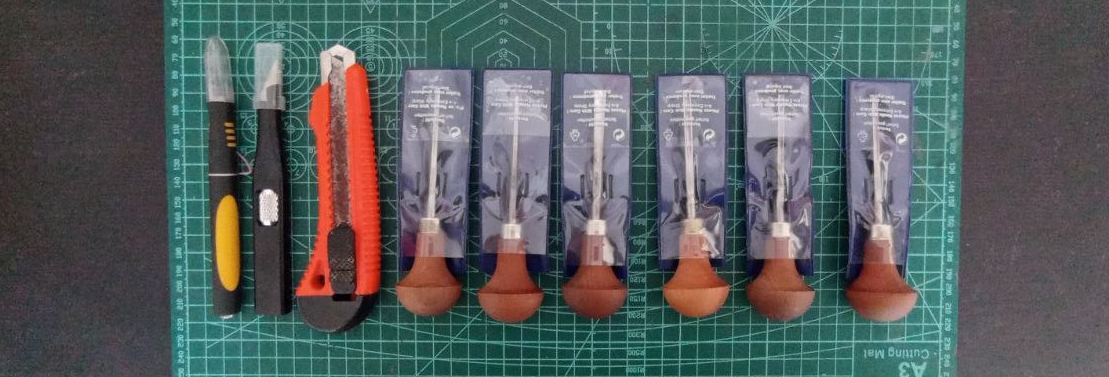Foto a una superficie de trabajo verde con varias gubias y cutters colocados encima, en fila, uno al lado del otro. Desde la izquierda: 2 cuchillas de modelismo de mango negro, un cuttter grande de cuerpo narajna, y seis gubias de mango redondeado de madera con los filos dentro de sus protectores de plástico azul y cubierta transparente. Todos los filos apuntan al borde superior de la imagen.