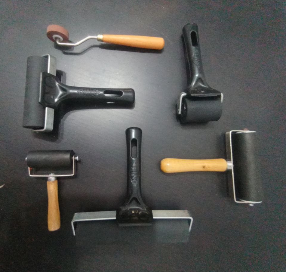 Foto a seis rodillos puestos sobre una superficie de madera negra veteada. Son de varias longitudes (de 1 a 20 cm). Todos con la goma del rodillo en color negro menos el pequeño, que es marrón. Tres tienen el mango de plástico negro, y 3 de madera clara. Los apliques del eje de los rodillos son todos de metal plateado. Están puestos de manera que tengan cierta armonía entre sí, pero cada uno colocado en una dirección distinta, como dentro de un cuadrado imaginario.