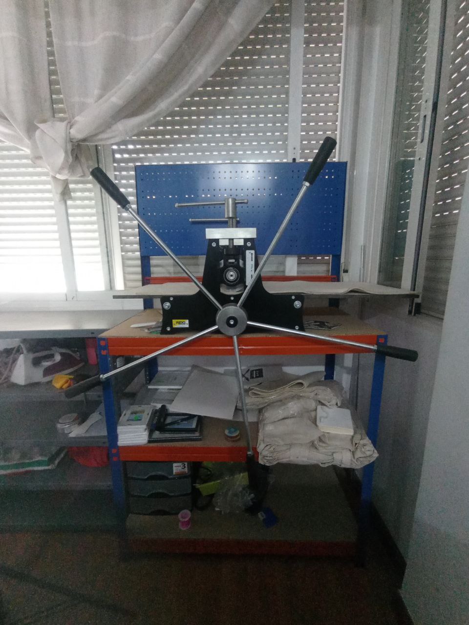 Foto a un tórculo con un aspa de cinco brazos. Está puesto sobre un banco de trabajo de metal azul y naranja. El banco está metido en una esquina entre unas ventanas con las persianas bajadas y con unas cortinas cogidas en un burruño para que no molesten. En las baldas inferiores del banco se ven cosas sin orden ni concierto (marcos, papeles, bolsas de tela, unos cajones de plástico... Al lado hay otro banco de trabajo de metal de color gris. Este no tiene nada encima, aunque en sus baldas inferiores también se ve lo manga por hombro que tengo todo (camisetas dobladas de varios colores, un trapo amarillo, una plancha, varios botes de tinta...).
