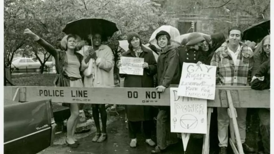 Foto en blanco y negro de varias personas tras una vaya policial en la que pone "POLICE LINE".
Las personas visten al estilo de los años 70 en Estados Unidos.
Aunque está lloviendo (a juzgar por varios paraguas que salen), su carácter es alegre, y sostienen varios carteles con consignas (que no consigo leer bien), pero que van a favor de los derechos para el colectivo gay.
En este grupo de gente ( en el que me parece contar 12 cabezas), a la izquierda del todo, se encuentran Sylvia Rivera y Marsha P. Jhonson
Sylvia aparece a la izquierda, con un vestido corto de color claro ceñido por un cinturón negro a la cintura, y lleva una chaqueta que parece cuero así como un bolso (ambos de colores oscuros). Aparece levantando el puño derecho en señal de protesta.
Marsha aparece a la derecha, con un chaquetón que parece de "pelo", con un bolso blanco. Sujeta un paraguas con el que resguarda a ambas. Tiene una flor en el pelo.
Esta es la foto en la que me baso para hacer el dibujo.