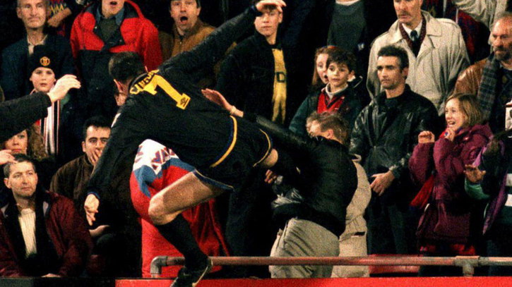 Foto apaisada de la foto de Cantona dando una patada en el pecho a un nazi. Cantona sale en el centro de la imagen, de espaldas, con botas de fútbol, calcetines, pantalones cortos y camiseta de manga larga, todo negro. Con detalles en dorado, como la palabra "CANTONA" (que a penas es legible por las arrugas de la camiseta) y el número 7. Tiene la pierna izquierda con casi ángulos rectos en ingle y rodilla; los brazos forman una T con el tronco del cuerpo; y la pierna derecha se apoya firmemente en el pecho del nazi que aparece a su derecha. El nazi lleva unos pantalones caquis y una cazadora negra y brillante, no se le ve la cara porque la tapa el brazo izquierdo que tiene levantado con la mano en alto (pero seguro que tiene una mueca de dolor). El pelo rubio lo lleva corto, pero no al 0. Algo menos de 20 personas salen junto al nazi, se ven expresiones de asombro.